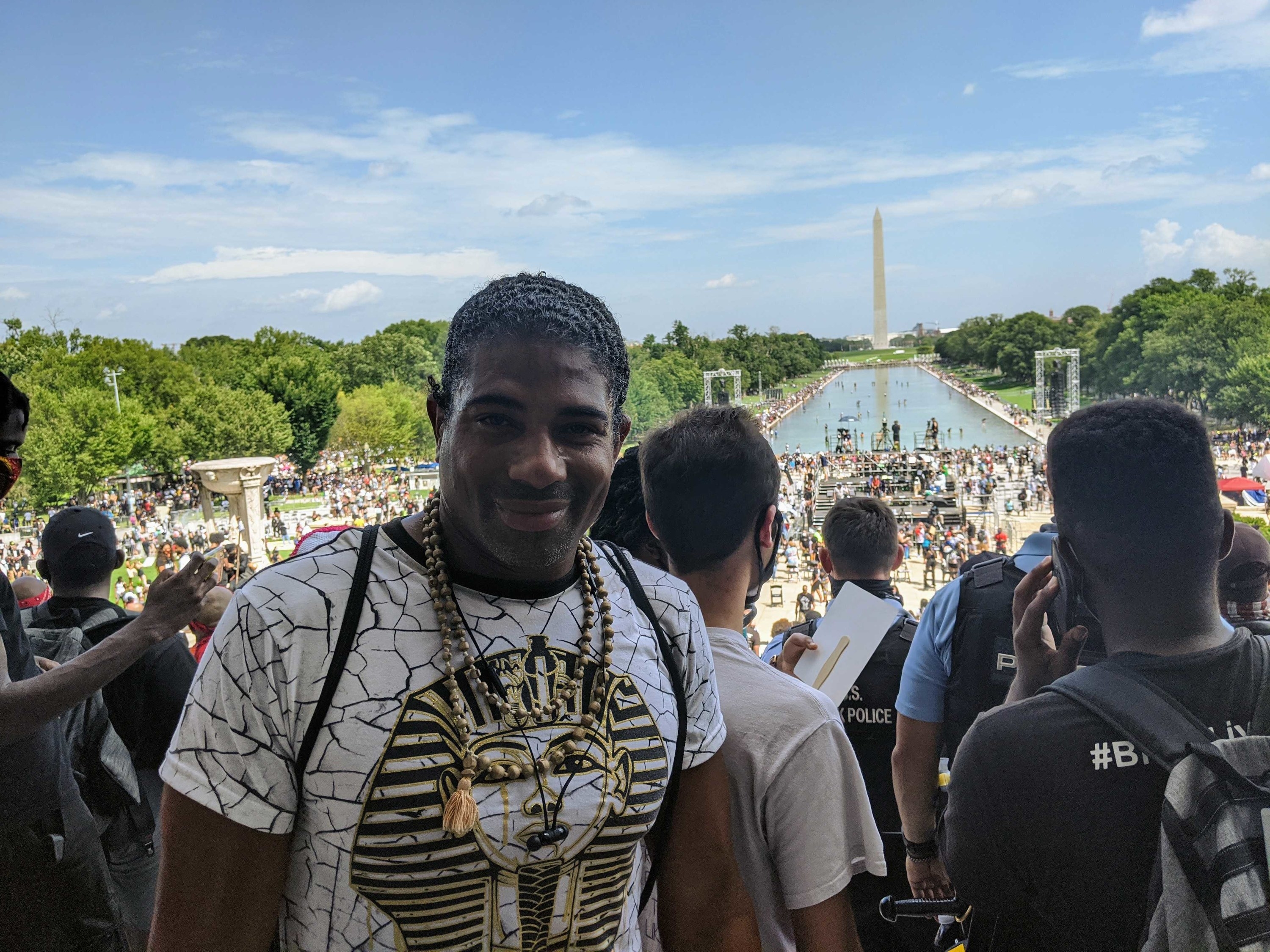 A man stands on the steps of the Lincoln Memorial with a crowd, the Reflecting Pool, and the Washington Monument behind him 