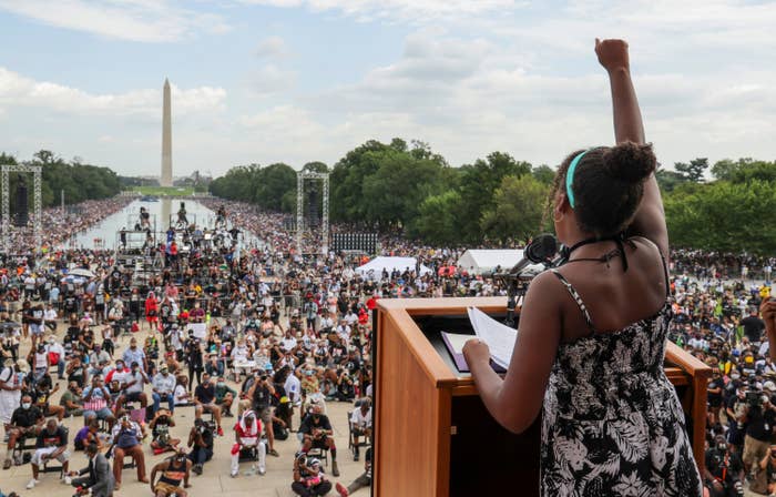 A young woman stands at a podium with her fist raised in the air in front of a large crowd leading down the Reflecting Pool to the Washington Monument