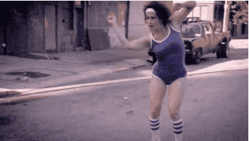 Ilana Glazer dancing as she rollerskates down the street in knee high socks, booty shorts, a tank top, and a sweatband 