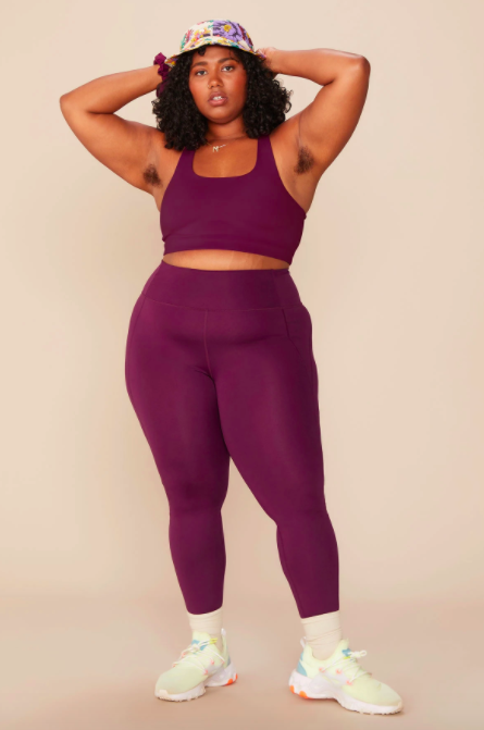Model wears berry-colored Girlfriend Collective high-rise leggings with a matching sports bra and colorful sneakers