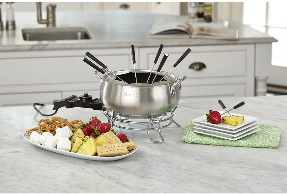 Fondue forks in a fondue bowl on a counter beside a plate of fruit and marshmallows