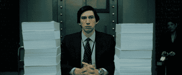 Adam Driver looks upset next to two stacks of paperwork in The Report