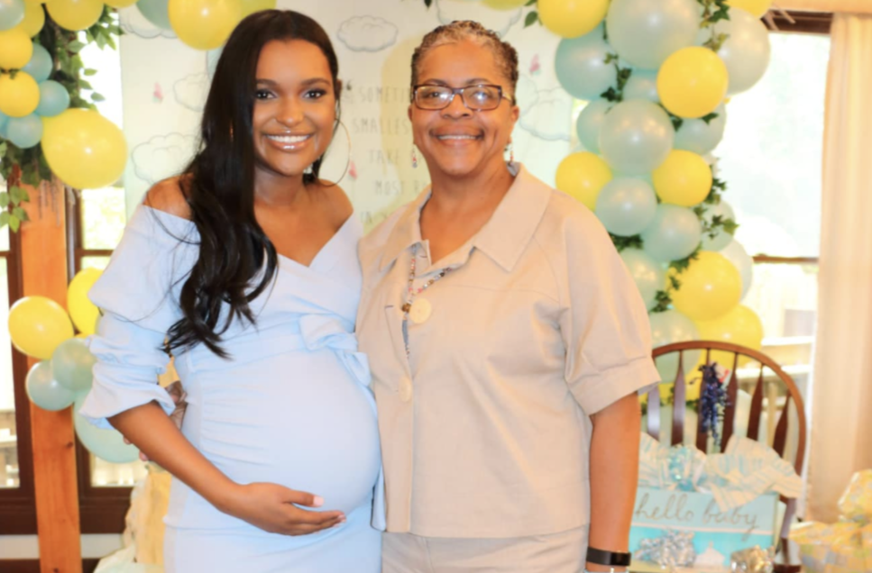 A pregnant Caitlyn Lewis holds her stomach while standing next to Kim Lewis Evans at a  baby shower with presents in the background