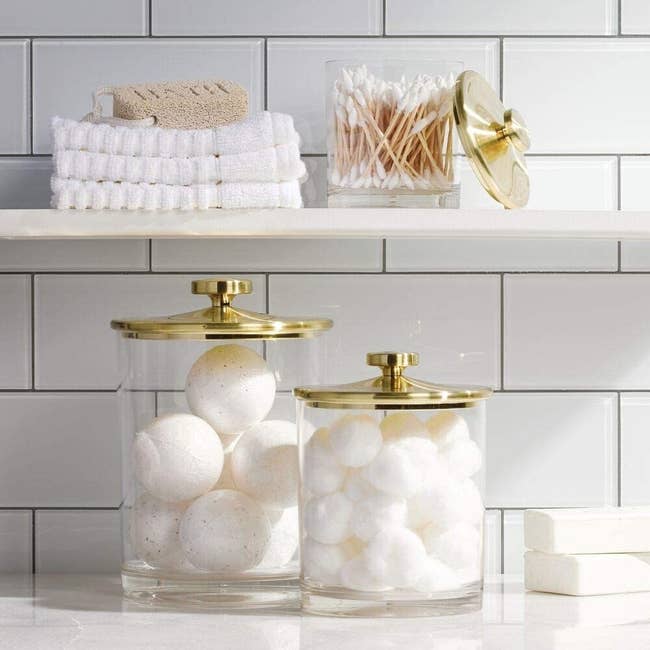A large and medium clear jar with a gold lid sitting on a counter with bath bombs and cotton balls in them and then a small jar on the shelf above with q-tips in it