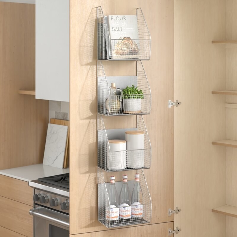 A shelving unit made of four metal baskets mounted on a cabinet door holding water bottles, jars, cookbooks, and a plant.