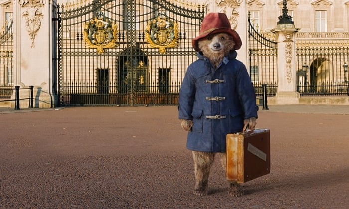 Paddington wearing a blue coat and red hat, standing in front of Buckingham Palace in London. He is holding a briefcase