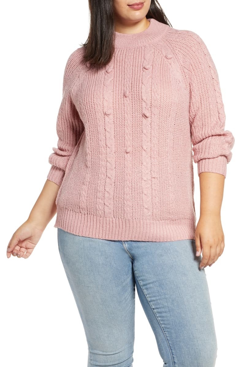 Model wearing Single Thread textured cable-knit sweater in pink with poms styled with light-wash jeans