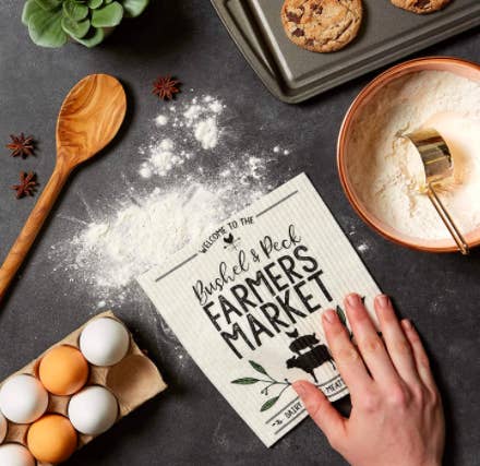 Hand uses reusable dishcloth that says &quot;Bushel &amp;amp; Peck Farmers Market&quot; to wipe up flour on a kitchen table