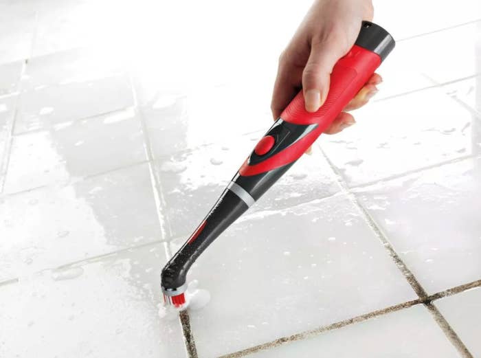 Hand holds red and black power scrubber cleaning grime from a shower tile