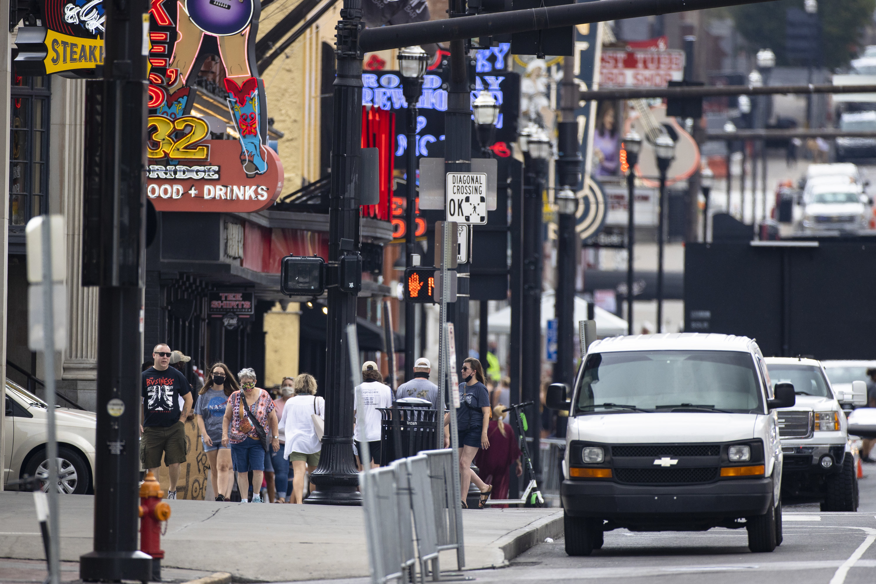 Visitors to Lower Broadway, some wearing masks, walk between businesses with illuminated signs advertising food and drinks