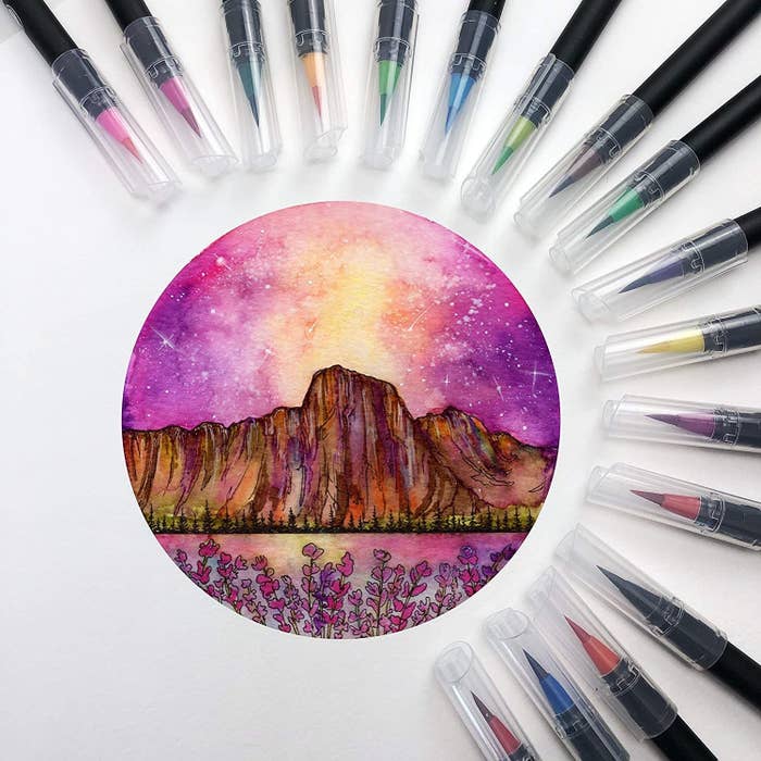 Twenty watercolour brush pens placed around a small drawing of a sun setting behind a mountain