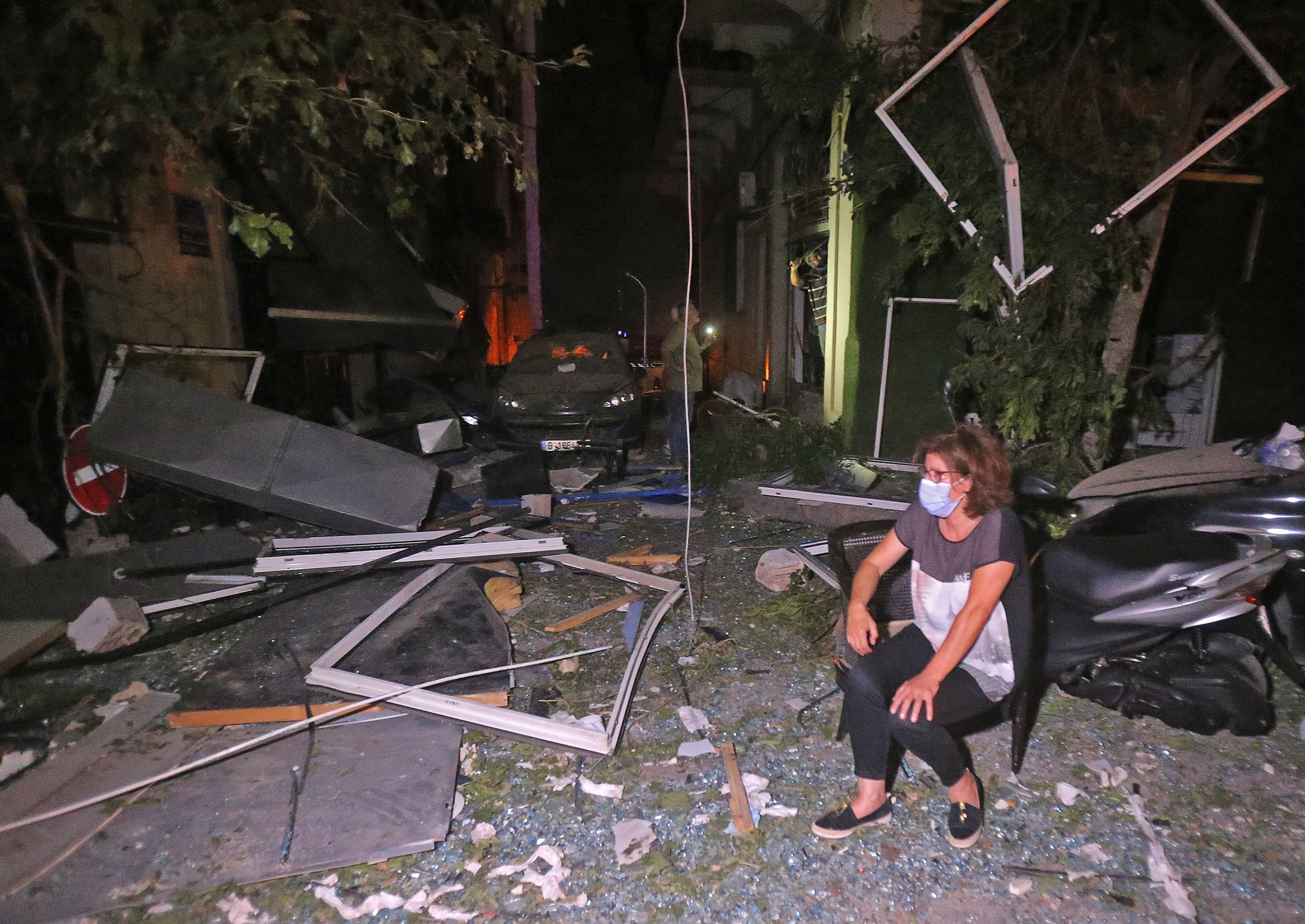 A woman sits on a chair at night on a street covered in glass and debris 