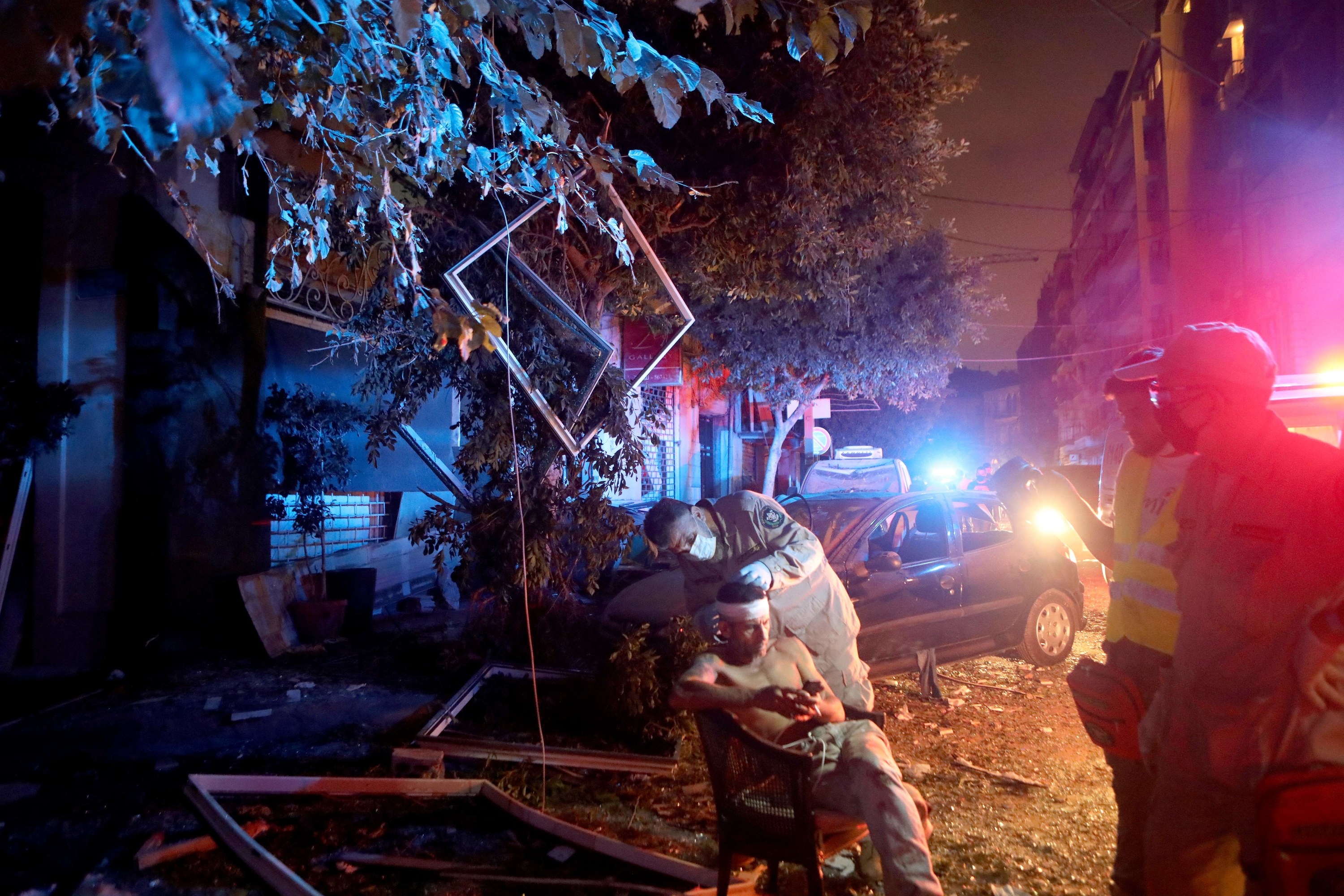 An injured man sits outside under red and white lights at night while a rescue worker examines his bandaged head