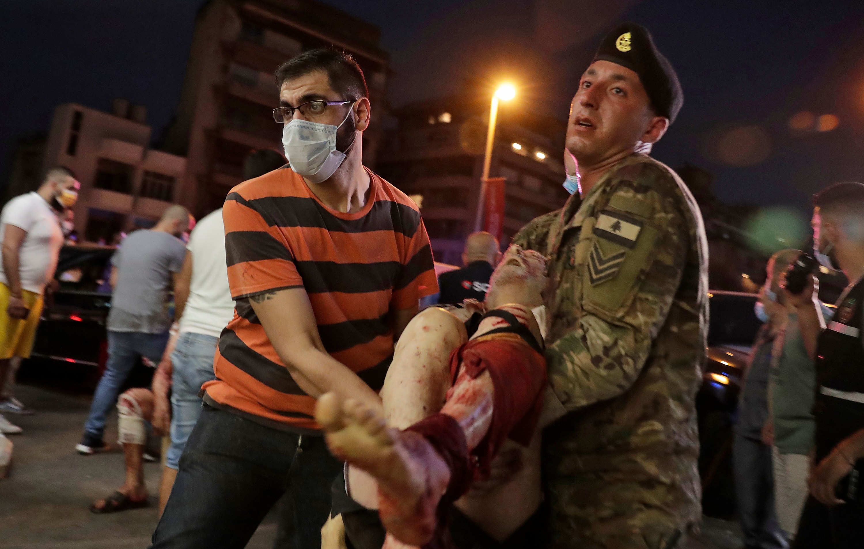 A man in a striped shirt and a mask carries a person with a leg injury down a crowded street with the help of a Lebanese soldier in uniform 