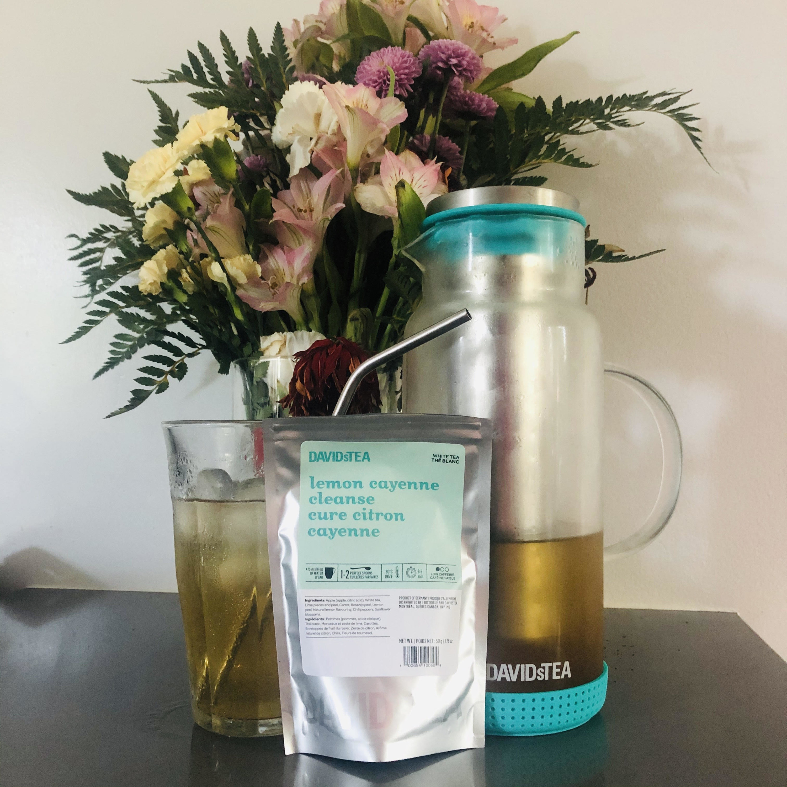 A bag of tea in front of a pitcher and glass of iced tea and a bouquet of flowers