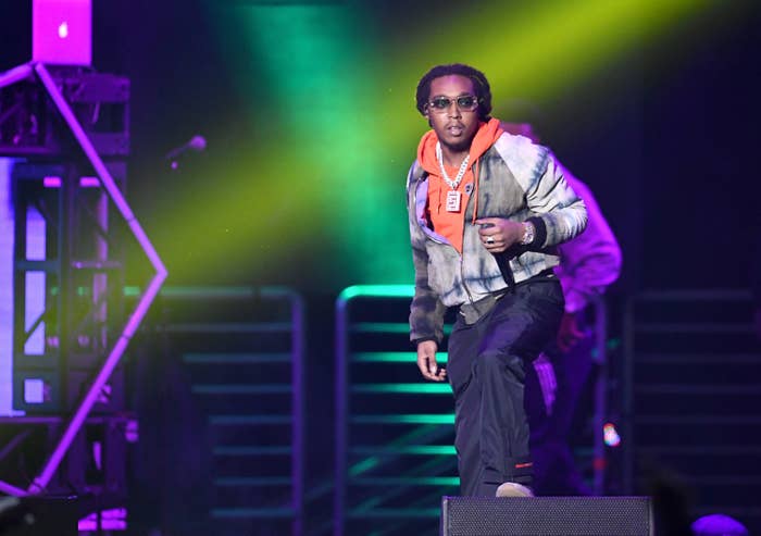 Takeoff stands onstage, wearing a hoodie, chain necklace, and jacket and holding a microphone