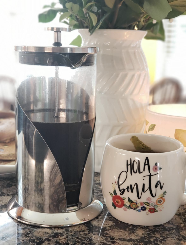 A reviewer image of the glass french press with stainless steel accents next to a cup of coffee 
