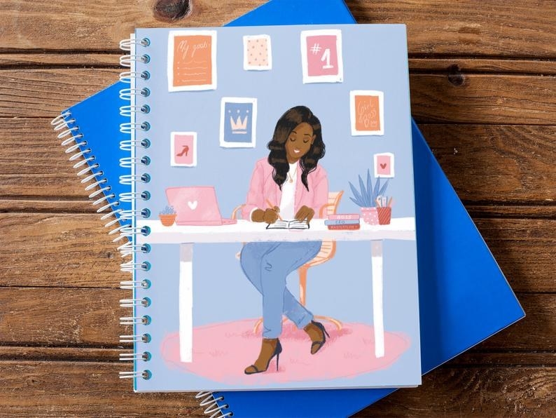 The 5.5x8.5 notebook with an illustration of a Black woman at her desk, wearing jeans, a pink blazer, and heels and surrounded by cute, empowering wall art