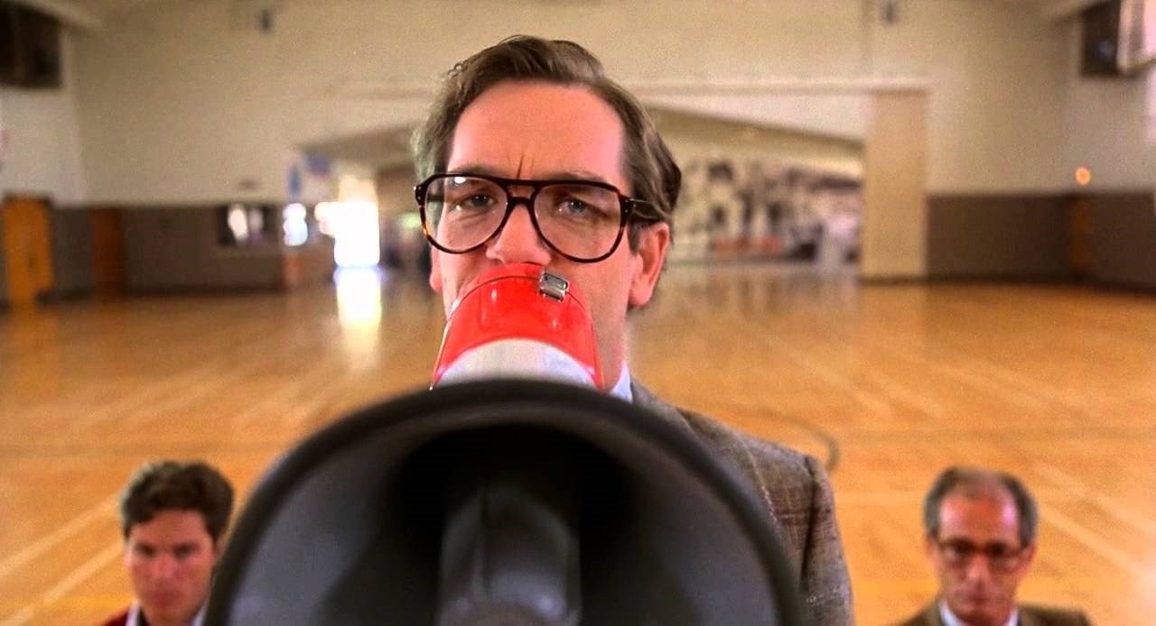 A straight-laced looking Huey Lewis holds up a bullhorn in a gymnasiun