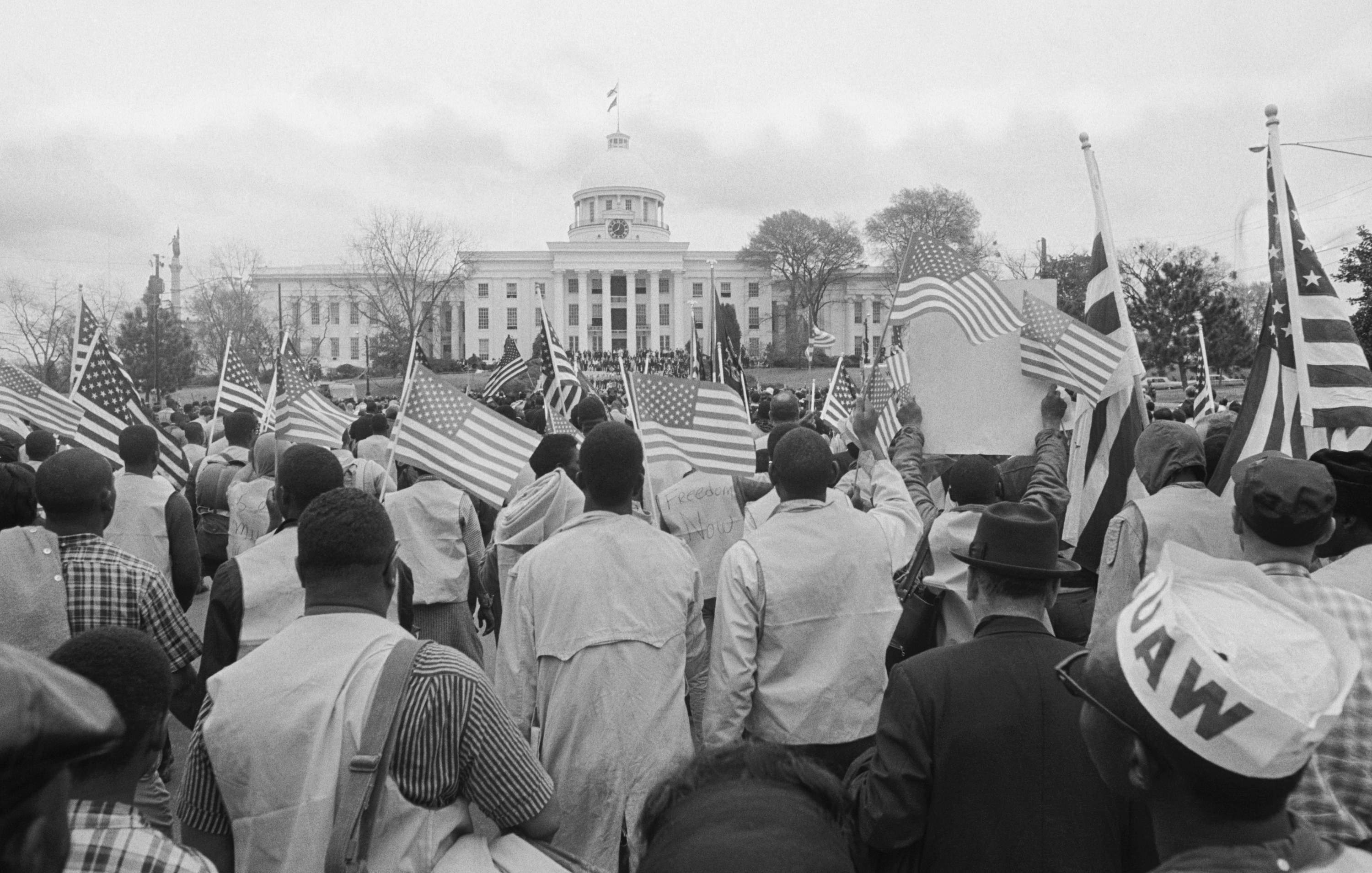 A crowd of black men and women carrying American flags are seen from behind as they stand in front of the capitol building in Alabama