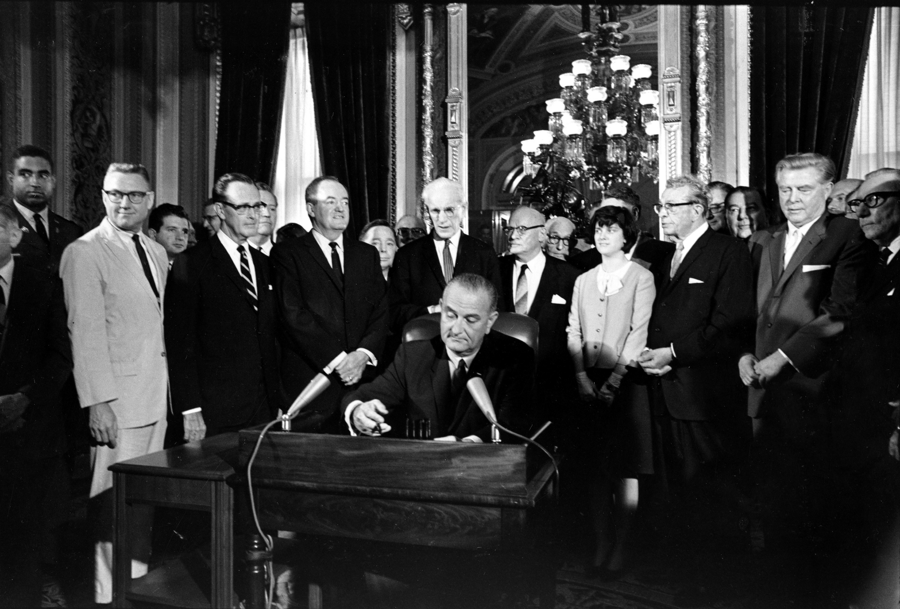 President Lyndon B. Johnson sits at a desk surrounded by white men in suits