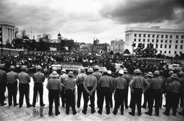 A line of police officers is seen from behind as they face a crowd in front of the capitol building in Alabama