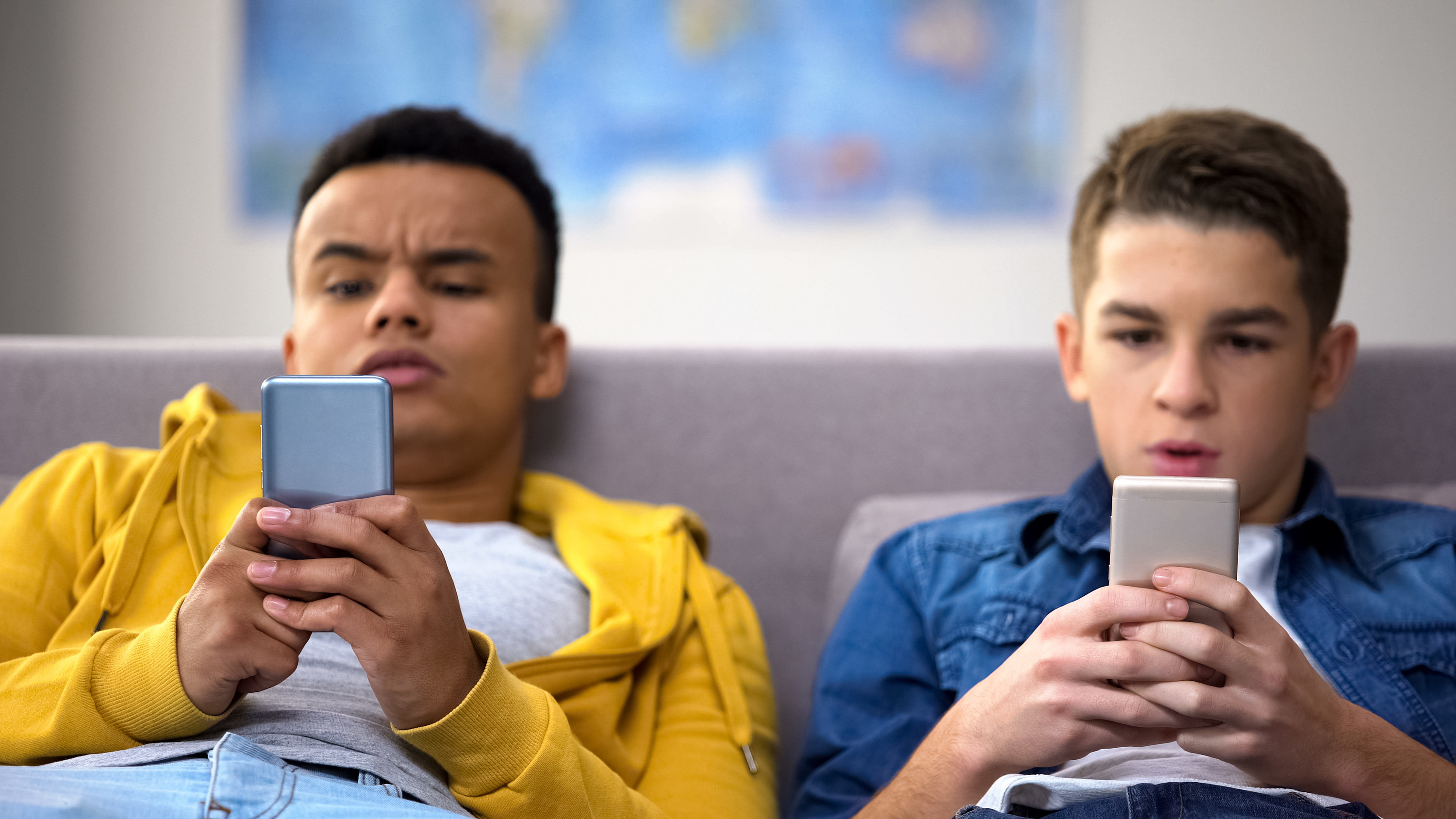 A stock image showing two young boys sitting on a grey couch on their phones