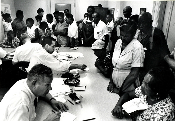 Black men and women stand at a table while white men sit behind it with registration papers. 