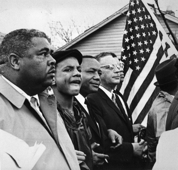 A group of five men, four black and one white, walk arm in arm down the street with an American flag