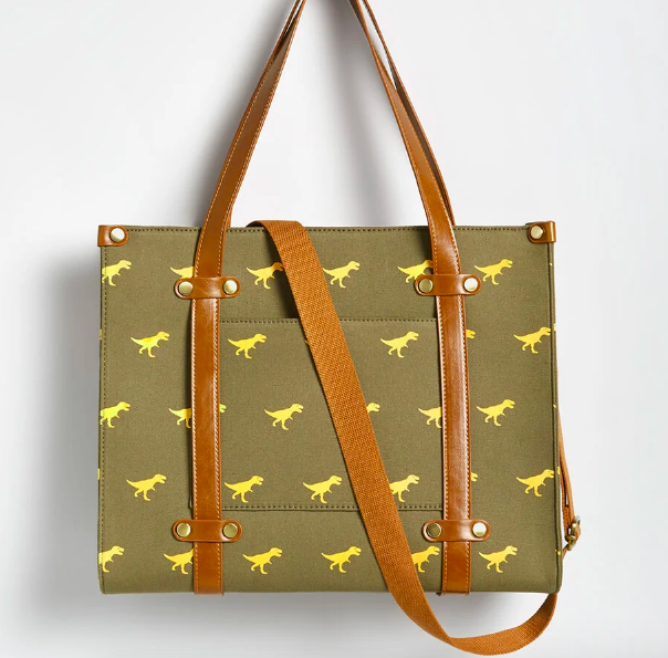 An olive green tote with warm faux-leather straps and gold button hardware. It has a pocket on the outside, a zipper at the top, and a tiny golden T-Rex pattern throughout the fabric.