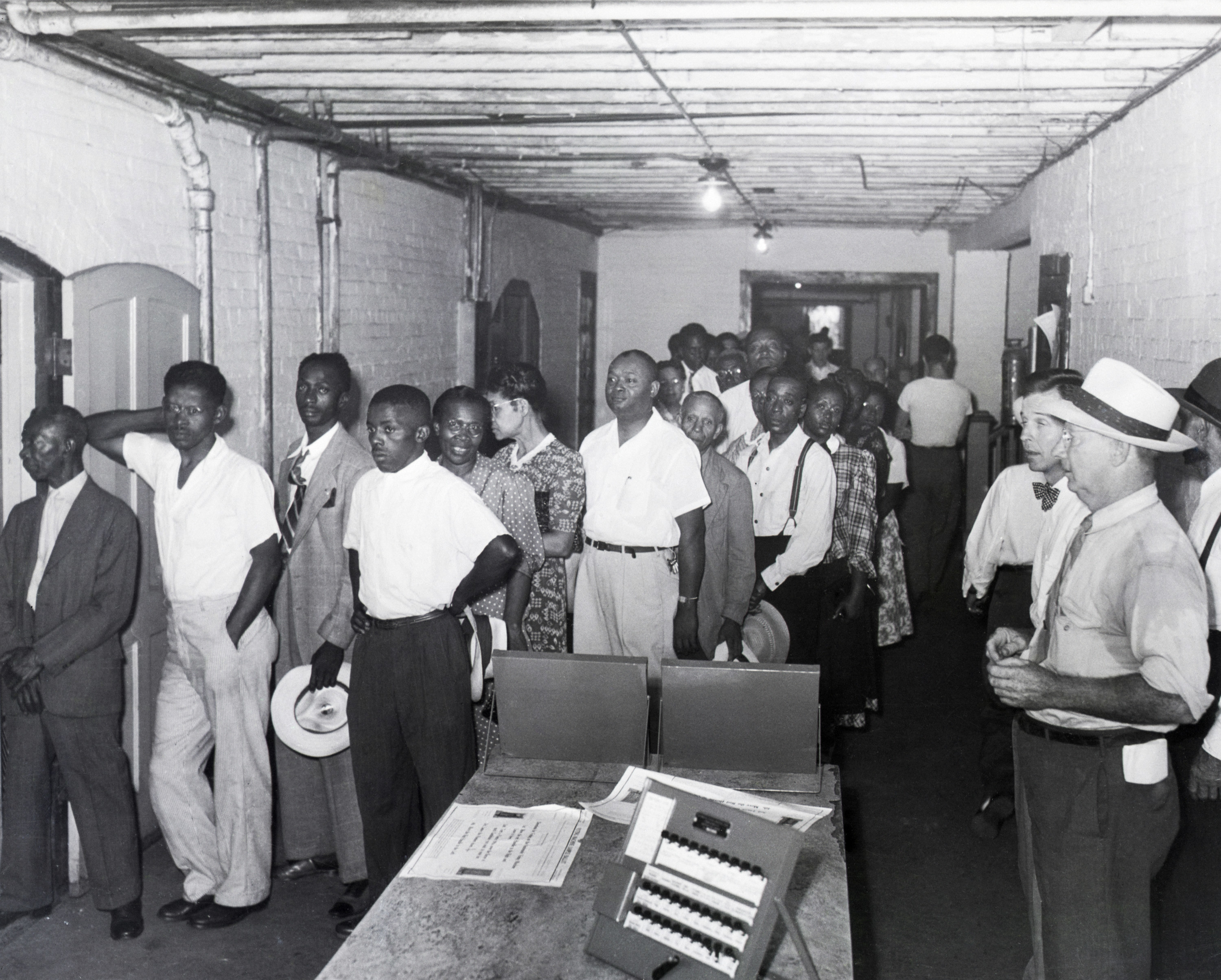 Black men line up, holding hats and dressed nicely, in a room where they are watched by white people