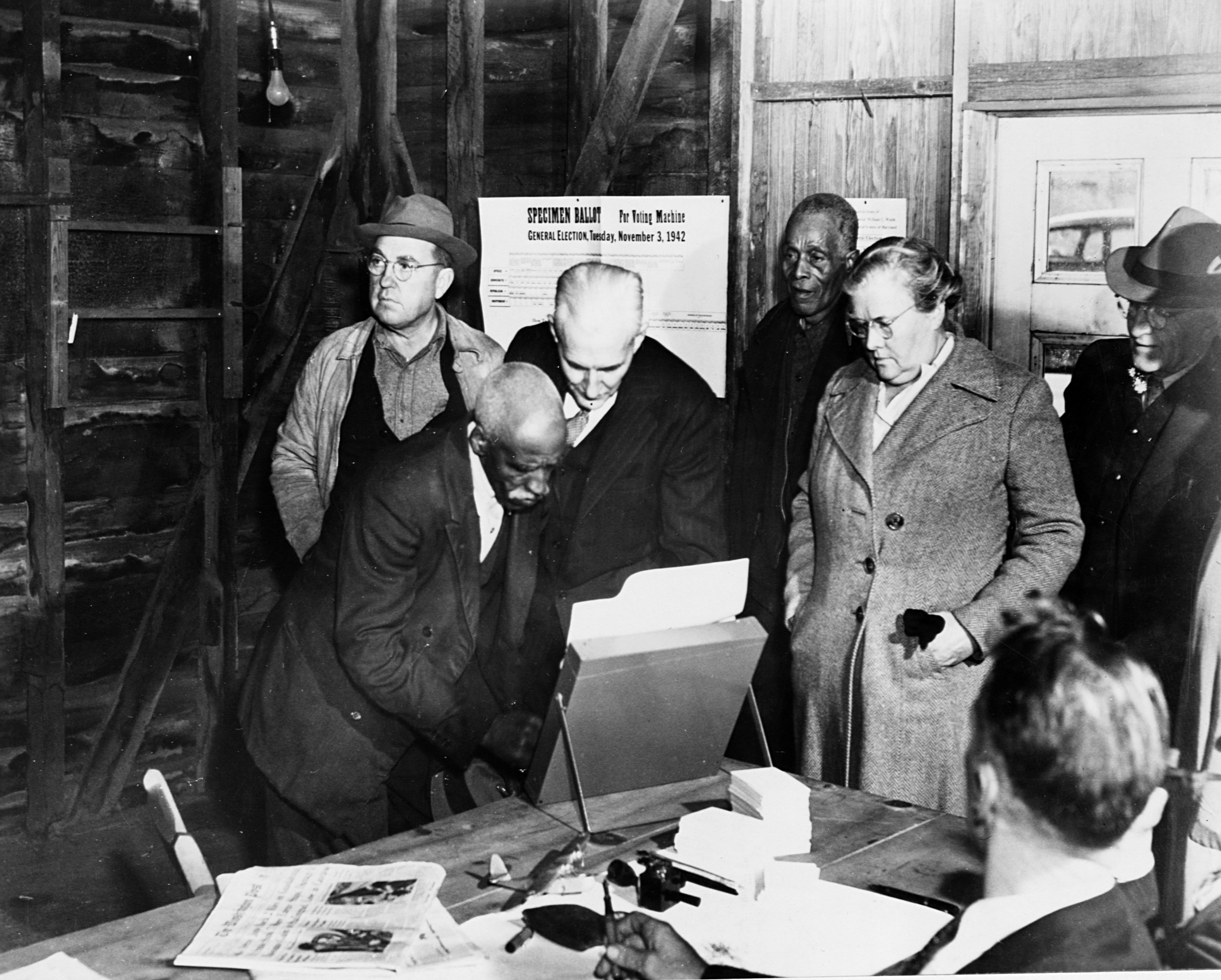 An elderly black man at the ballot box while a line of white and black people in overcoats wait behind him