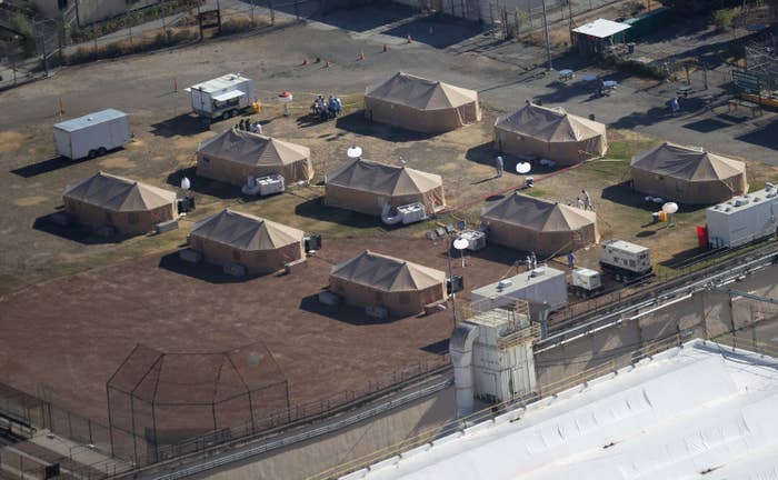 Nine large, tan tents stand in an empty field surrounded by fencing and barbed wire