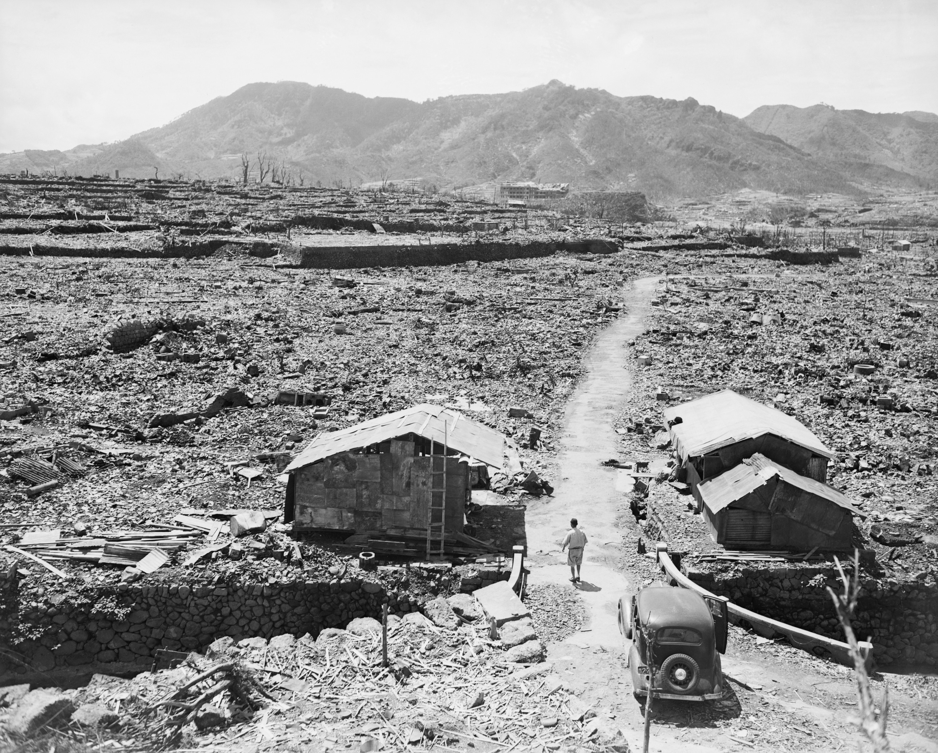 A man walks away from a car and toward a lone building standing amid rubble in a destroyed valley