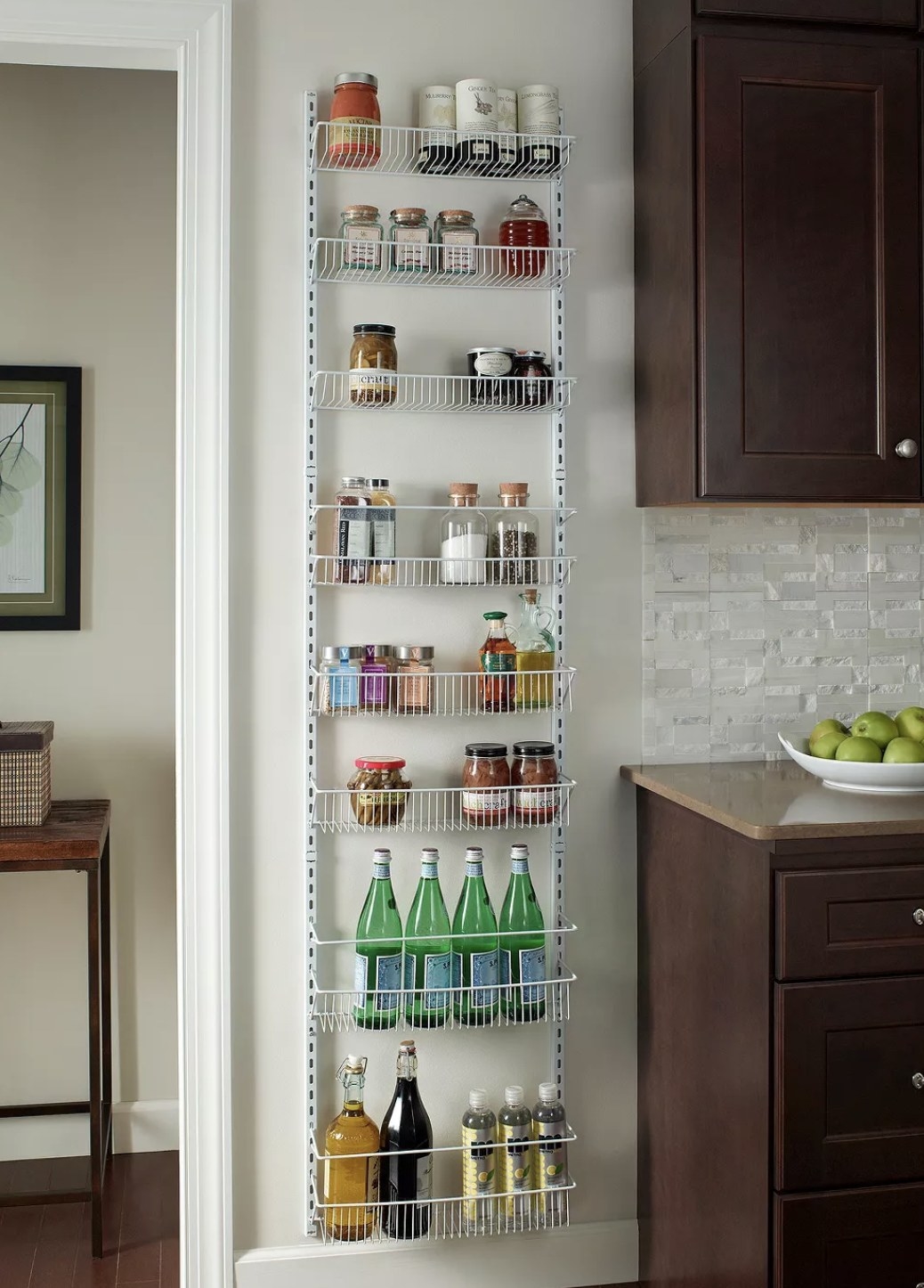 A wire rack mounted on the wall of a kitchen holding various spices, oils, sauces, and large seltzer bottles