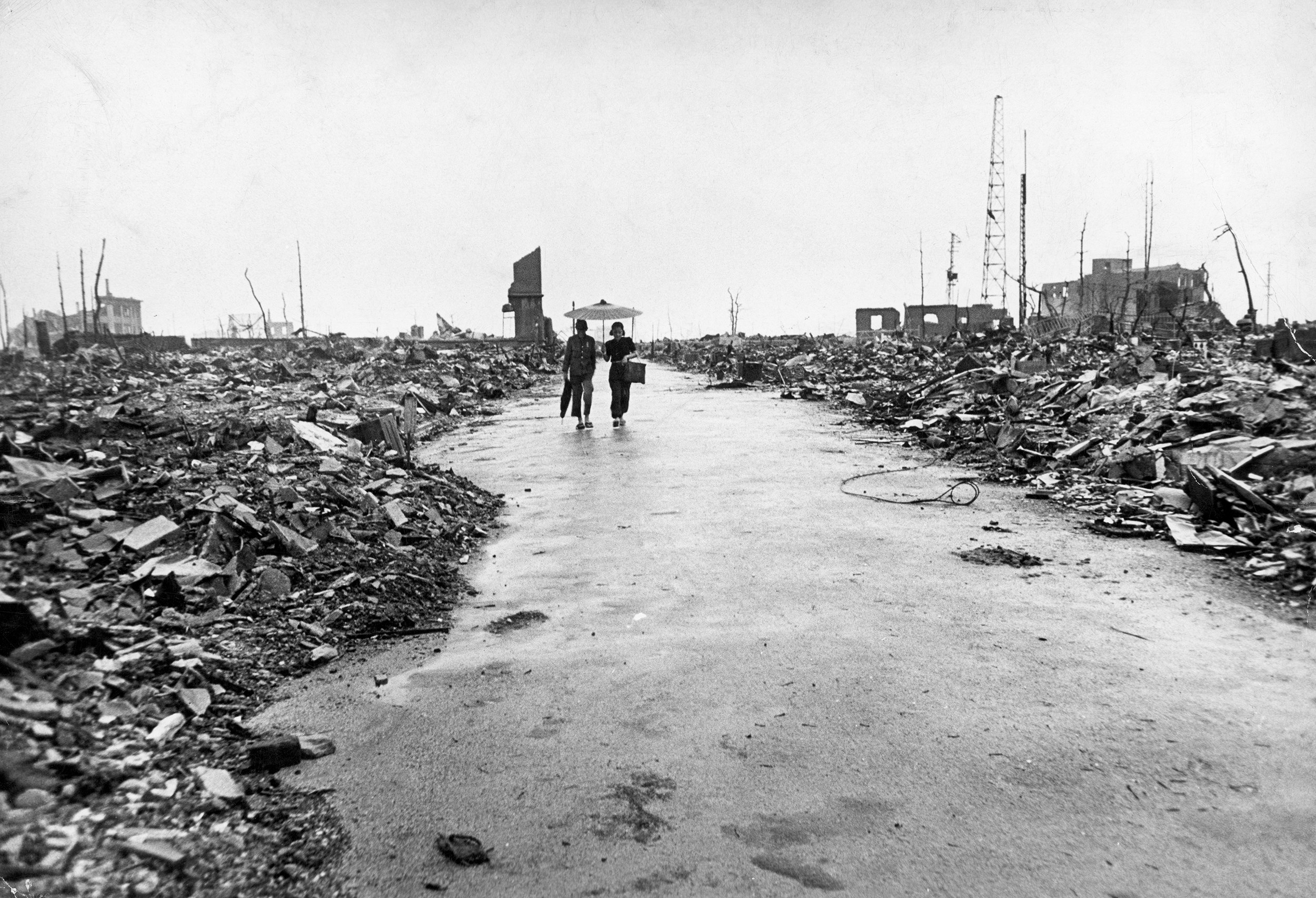 Two people, one carrying an umbrella, walk down a street of rubble. 