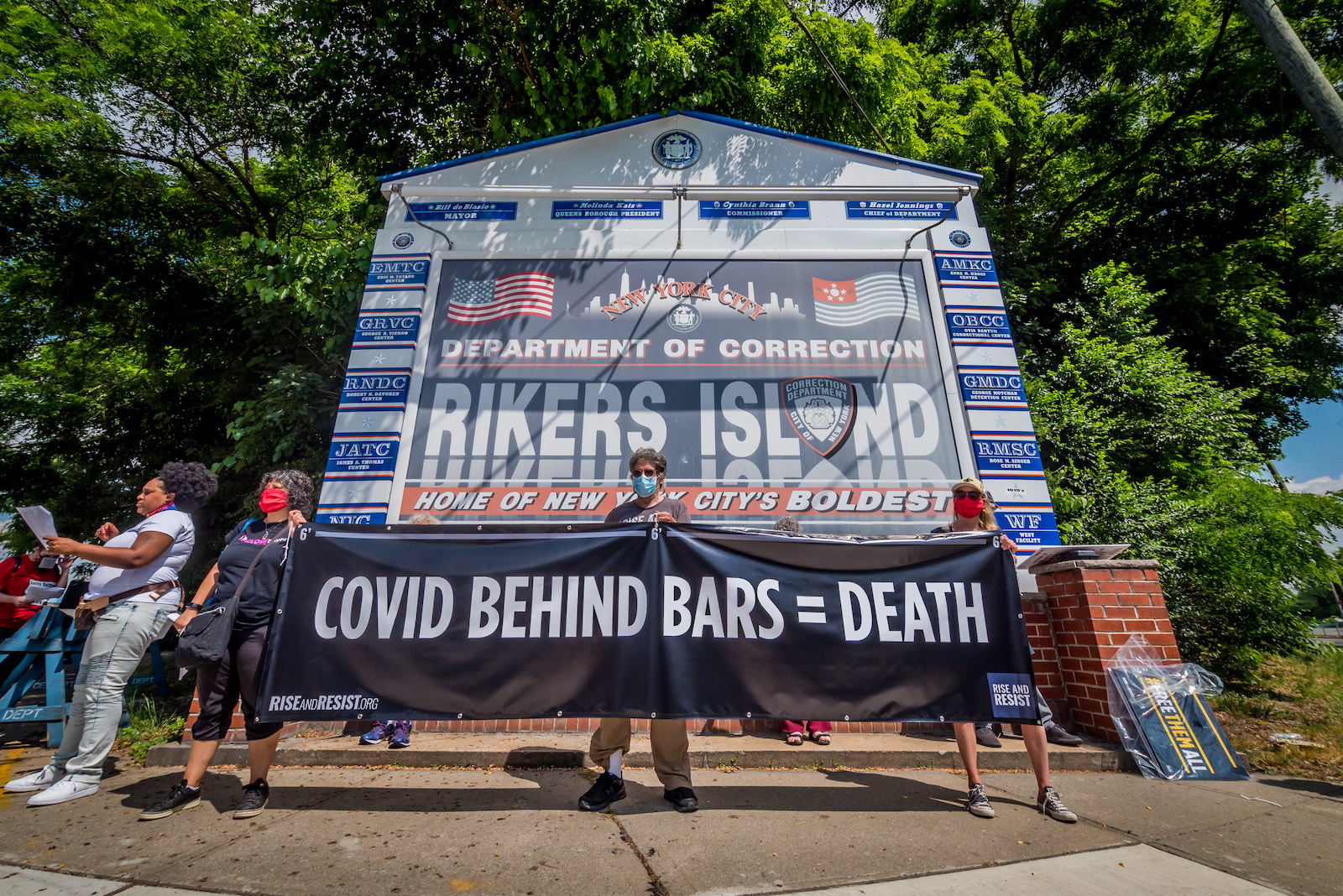 People in masks hold up a banner reading &quot;COVID BEHIND BARS = DEATH&quot; in front of the sign for Rikers Island