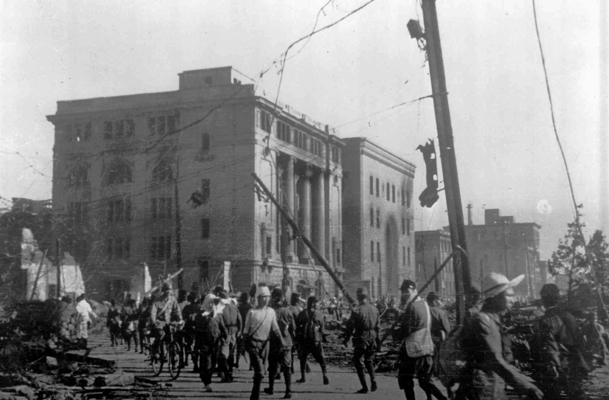 A crowd of mostly men, some in soldiers uniforms, walk down a city street surrounded by destroyed buildings