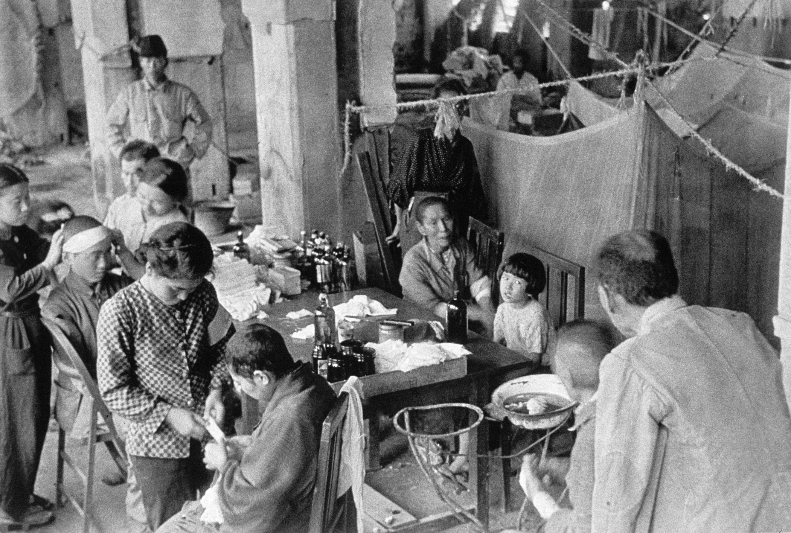 Children and people with bandages sit at a cluttered table and are tended to by women and older men