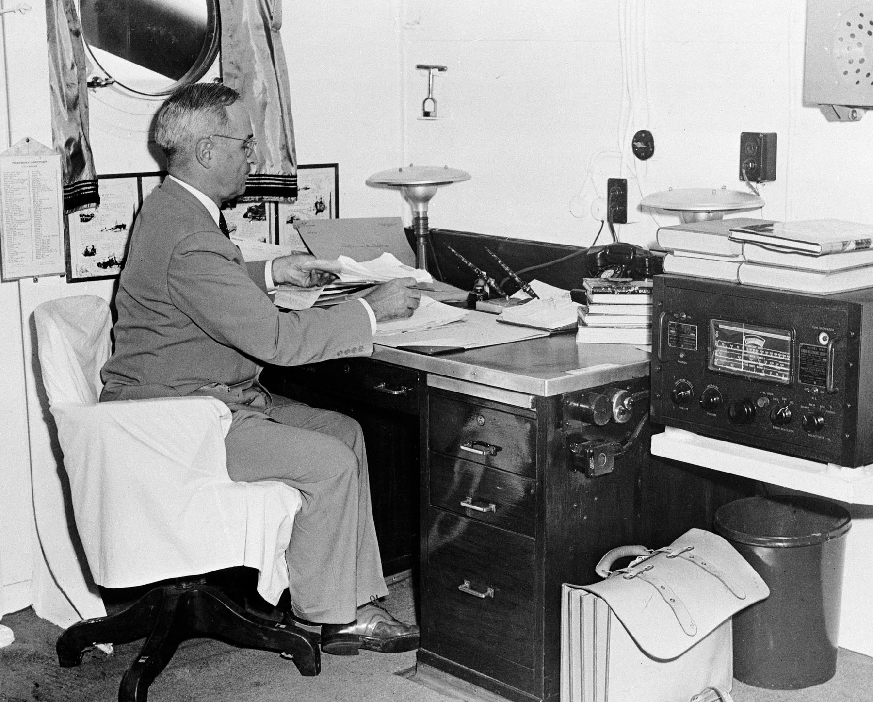 President Harry Truman sits at a desk with papers in front of him and a radio next to him 