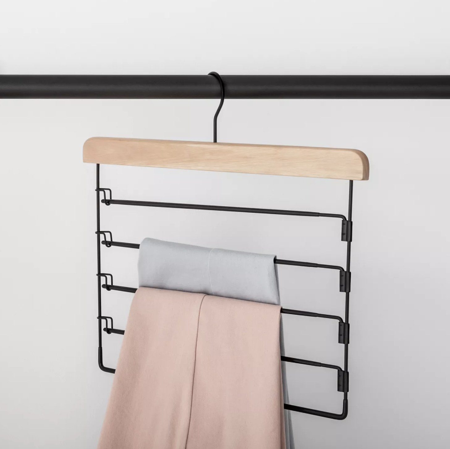 A wooden hanger with five black rungs holding a pair of blue pants and a pair of pink pants on a black closet rod