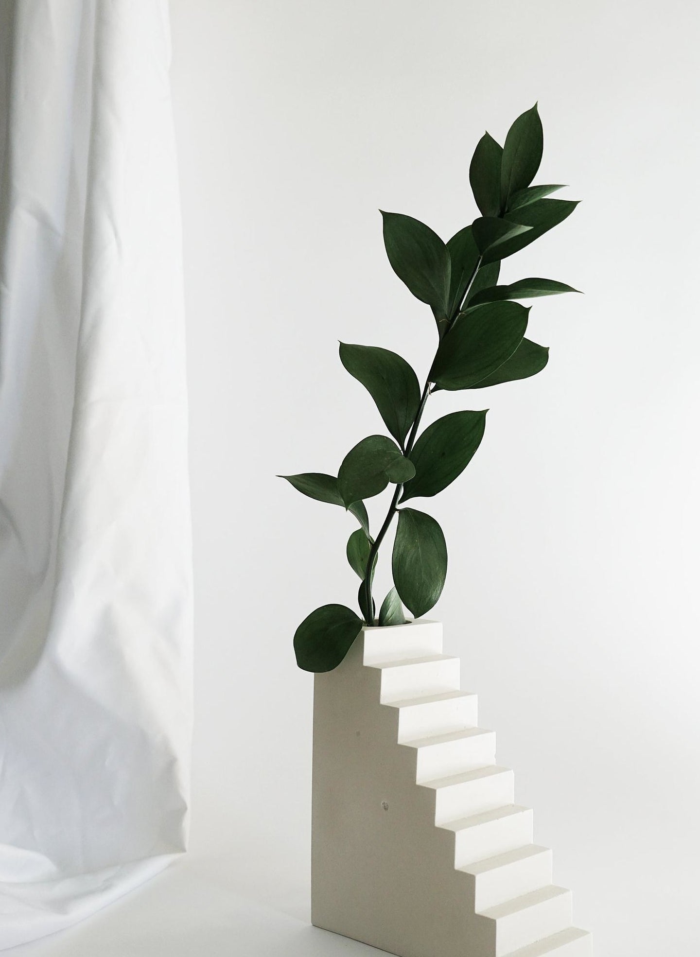white concrete vase shaped like a flight of stairs with a small rectangular opening on top holding a leafy branch