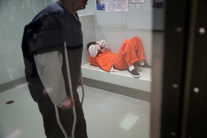 Two detainees are seen through a glass window, one in an orange jumpsuit lying down and another in blue jumpsuit standing with crutches
