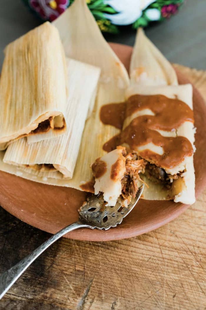 A rustic plate holds an old-fashioned spoon and a pile of tamales topped with a rich mole sauce as shreds of moist chicken peek out beneath the masa.