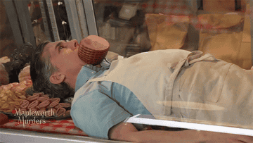 A dead Ben Canelli, played by Chris Parnell, with a large ham shoved in his mouth, The GIFs title says &quot;murder by ham.&quot;
