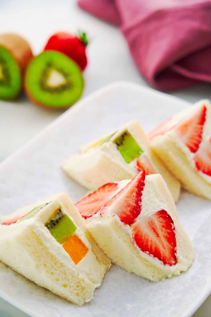 Four sandwiches made of soft white bread, fluffy whipped cream, and fresh slices of fruit