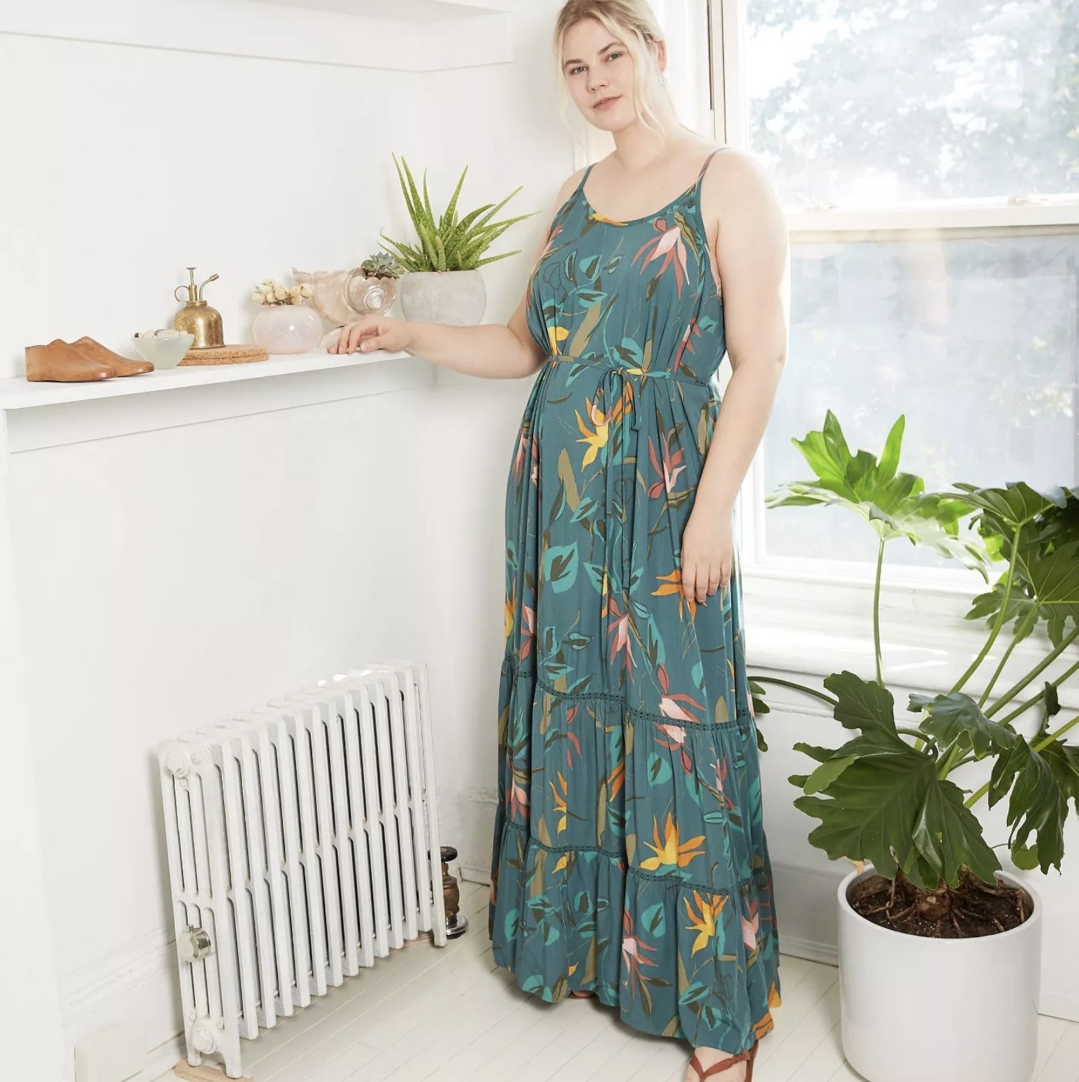 Model is wearing a blue/green maxi dress with thin shoulder straps and a floral pattern