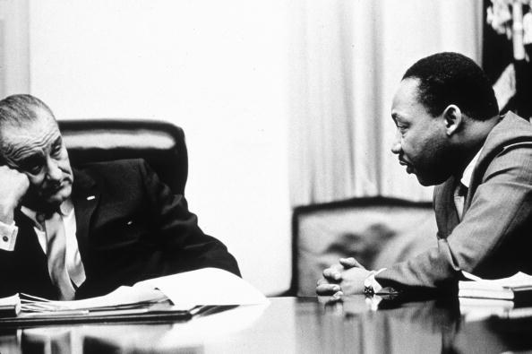 President Johnson is seen with his head leaning on his hand sitting at a table with a stack of papers in front of him while Martin Luther King Jr with his hands folded leans in to talk to him 