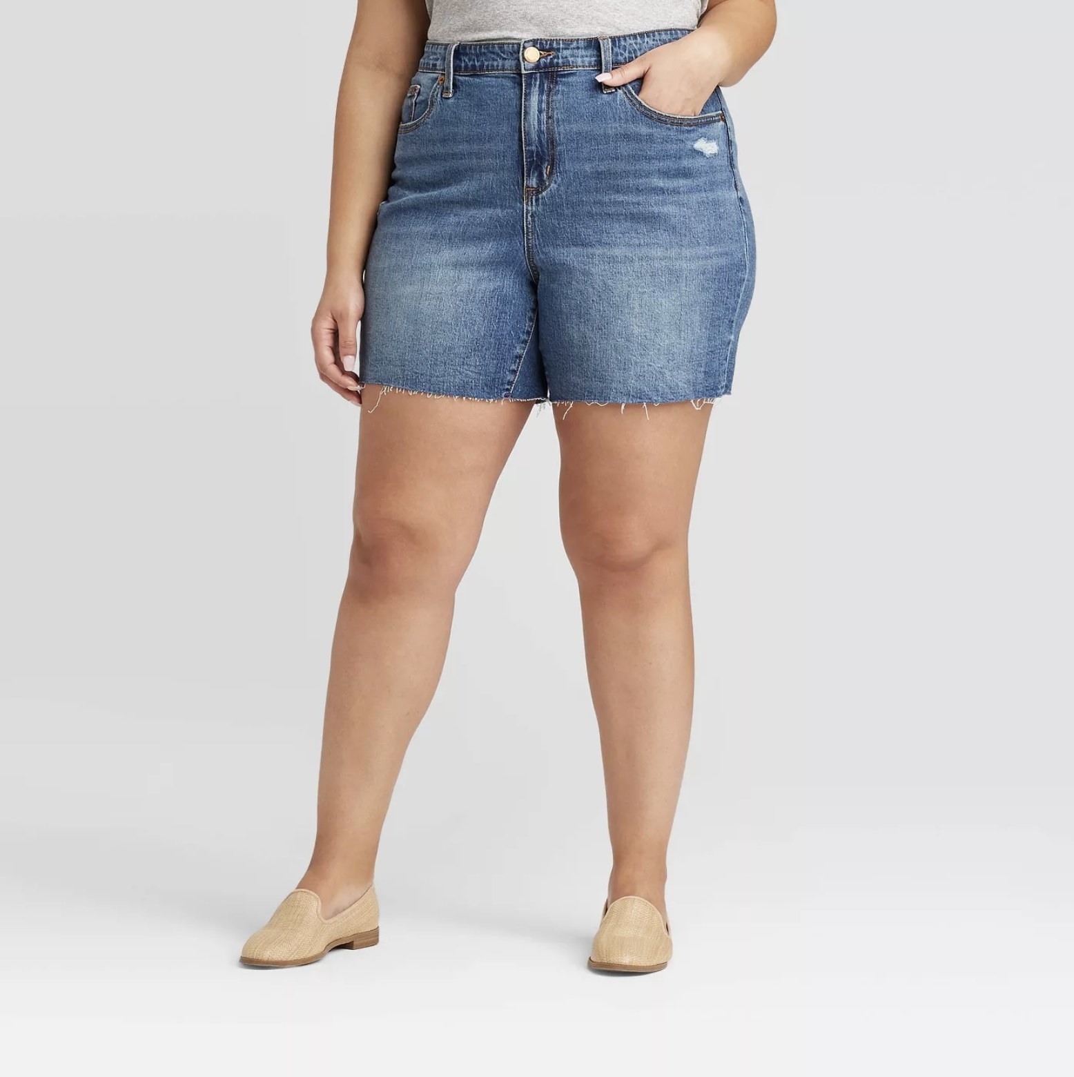 Model is wearing mid-thigh, high-waisted denim shorts in a classic denim blue with distressed hemline and light wash on the thighs