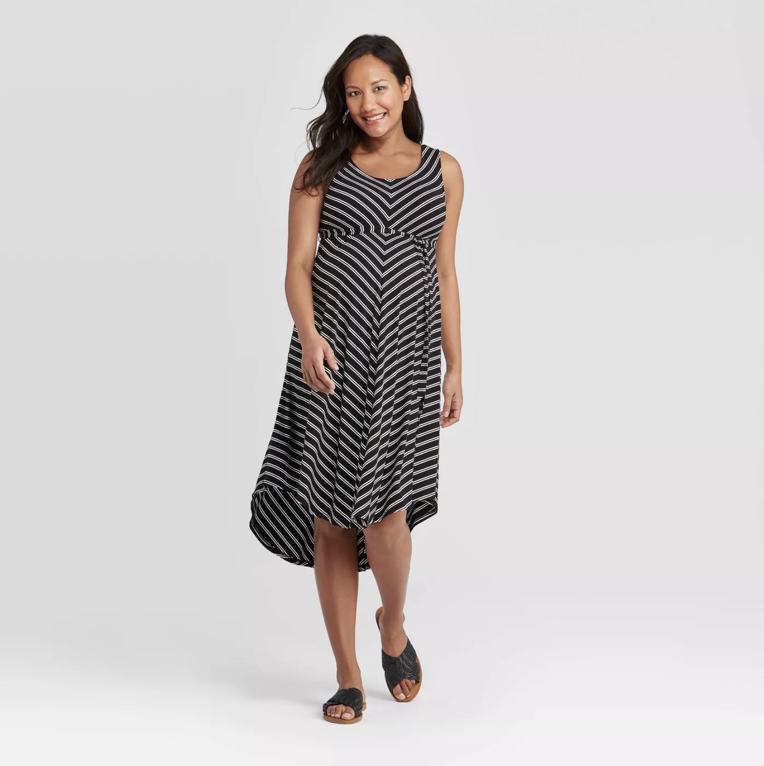 Model is wearing a black and gray striped maternity dress with thick shoulder straps, an irregular hemline, and a pair of black sandals